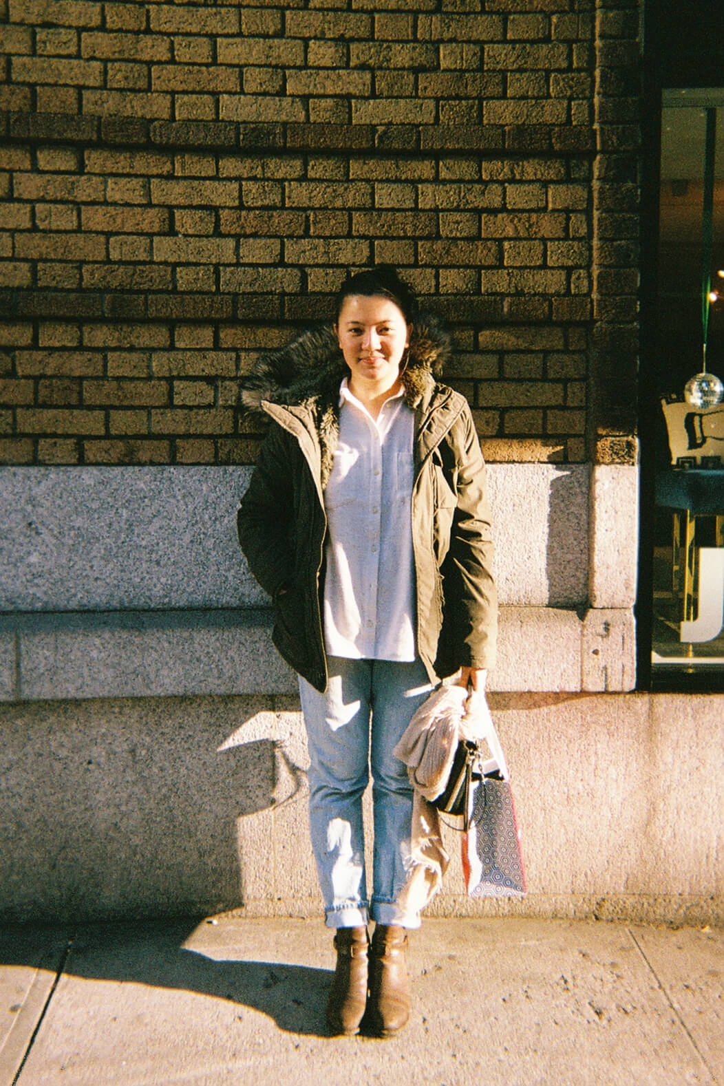 Full body shot of Gabriela Kresentia Ong in a white shirt, blue jeans and an olive green coat, front of a brick wall.